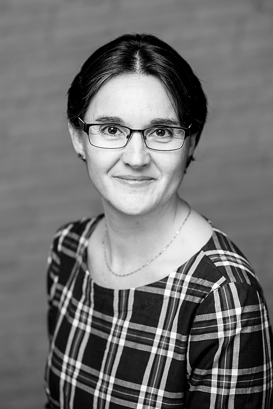 Schwarz-Weiß-Porträt einer freundlich lächelnden Frau mittleren Alters mit kurzen dunklen Haaren, rechteckiger Brille und einem karierten Oberteil. Sie strahlt Professionalität und Zugänglichkeit aus.